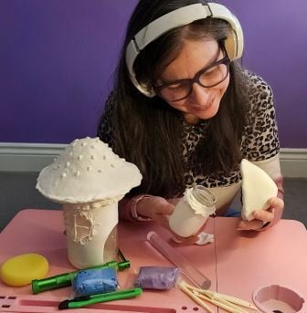  Canal Crafty Corner provides adult creative group workshops and sensory-friendly drop-ins, which the woman, pictured, is attending. She is using a personal workstation with natural light and listening to music with her headphones on while creating a fairy garden house out of clay.